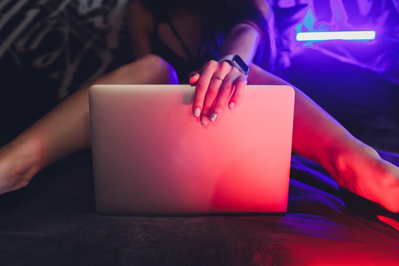 Closed laptop lit by red and purple neon in a dark bedroom